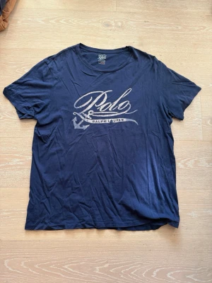 Mörkblå Polo Ralph Lauren t-shirt - Snygg mörkblå t-shirt från Polo Ralph Lauren med stort tryck framtill i vitt och grått. Klassisk rund halsringning och korta ärmar. Tillverkad i mjuk bomull som känns skön mot huden. Perfekt för en avslappnad och stilren look.