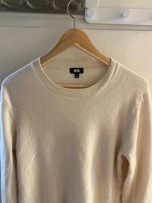 Beige stickad tröja från Uniqlo - Säljer en stilren beige stickad tröja från Uniqlo. Tröjan har rund halsringning och ribbstickade muddar vid hals, ärmslut och nederkant. Perfekt för lager-på-lager och enkel att matcha med olika outfits.