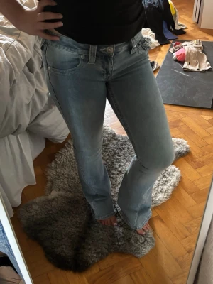 Ljusblå lågmidjade bootcut jeans  - Säljer ett par ljusblå bootcut jeans med snygga slitningar på bakfickorna. Jeansen har klassisk femficksdesign, låg midja och är i stretchigt denimtyg som sitter skönt. Perfekta för en avslappnad och trendig look. 