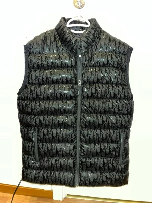 Svart dunväst från Dior - Snygg svart dunväst från Dior med glansig finish och ikoniskt monogrammönster över hela västen. Västen har hög krage, två dragkedjefickor och stängs med dragkedja framtill. Perfekt för lager-på-lager och streetstyle.