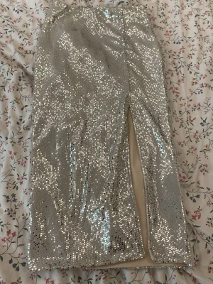 Glittrig silverkjol med slits - Säljer en glittrig silverfärgad kjol med paljetter och en snygg slits framtill. Kjolen är rak i modellen och har en festlig look som verkligen sticker ut. Perfekt för dig som vill glänsa lite extra.