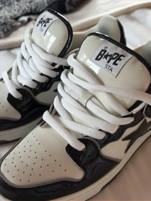 Svarta och vita BAPE STA sneakers SK8 version (sällsynta) - Säljer ett par BAPE STA sneakers i svart och vitt med glansig finish. Tjock plös för en mer relaxed look. Skorna har klassisk låg siluett, vita skosnören och tydlig BAPE-logga på plösen och innersulan. Materialet är lackat läder som ger en clean och modern vibe. 
