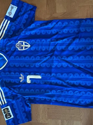 Blå retro fotbollströja Adidas - Säljer en blå retro fotbollströja från Adidas med vita detaljer och mönstrad design. Tröjan har klassiska tre ränder på axlarna, broderad klubbmärke och nummer 1 på bröstet. Tillverkad i lätt och ventilerande syntetmaterial, perfekt för match eller träning.