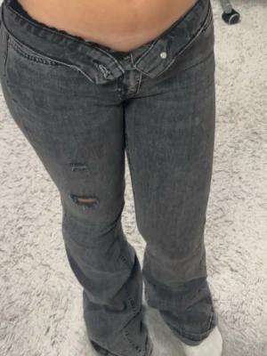 Grå bootcut jeans - Säljer ett par grå highwaist bootcut jeans och slitningar på ena benet. Jeansen har klassisk femficksdesign och stängs med knapp och dragkedja. Perfekta för en avslappnad och trendig look.