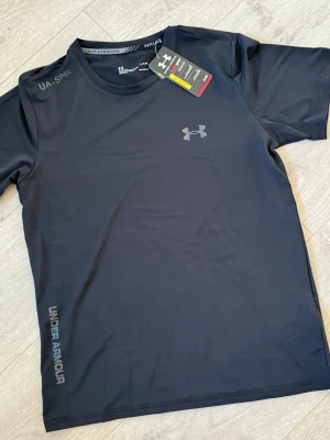 Svart tränings-tshirt Under Armour - Snygg svart tränings-tshirt från Under Armour med diskret logga på bröstet och text längs sidan. Tillverkad i lätt och snabbtorkande material, perfekt för löpning eller gym. Rund halsringning och korta ärmar för maximal rörelsefrihet.