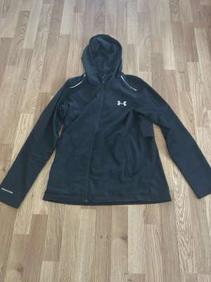 Svart träningsjacka från Under Armour - Svart träningsjacka med huva från Under Armour. Jackan har dragkedja framtill, två fickor och reflekterande detaljer vid axlarna. Tillverkad i lätt syntetmaterial, perfekt för utomhusaktiviteter.