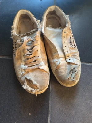 Saint Laurent court - Saint Laurent sneakers i vitt med tydliga slitningar och trasiga detaljer. Låg modell med snörning och handskriven logga på sidan. Tillverkade i canvas och läder, med vintage-inspirerad look och råa kanter för en edgy stil.