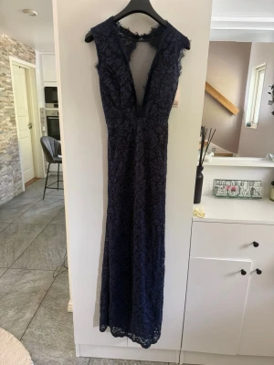 Marinblå långklänning med spets - Elegant marinblå långklänning med djup v-ringning och spetsdetaljer. Klänningen är ärmlös och har en figurnära passform med vacker spets över hela plagget. Perfekt för speciella tillfällen när du vill sticka ut. Helt ny, prislapp kvar