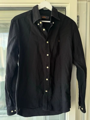 Svart skjorta från Morris - Klassisk svart skjorta från Morris med vita knappar och broderad logga på bröstet. Skjortan har lång ärm, normal passform och är tillverkad i mjuk bomull. Perfekt för en stilren look.