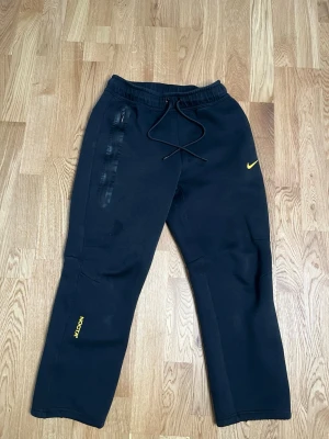 Nocta tech Nike svart set - Svart Nike Tech Fleece set med byxa och hoodie. Båda plaggen har dragkedjeförsedda fickor med svarta detaljer och Nike-logga i gult. Hoodien har hel dragkedja och huva. Byxan har resår och dragsko i midjan samt NOCTA-tryck i gult på benet.