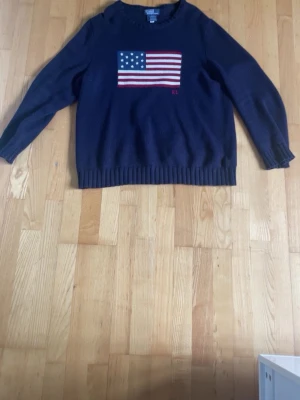 Mörkblå stickad tröja Ralph Lauren - Mörkblå stickad tröja från Ralph Lauren med amerikansk flagga i rött, vitt och blått på bröstet samt RL-broderi. Tröjan har rund halsringning, långa ärmar och ribbstickade muddar vid ärmslut och nederkant. Perfekt för en klassisk och avslappnad stil. Ny pris :5000 kr.
