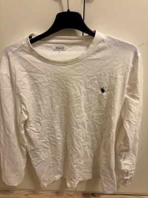 Vit långärmad topp från Polo Ralph Lauren - Klassisk vit långärmad topp från Polo Ralph Lauren i mjuk bomull. Enkel design med rund halsringning och den ikoniska lilla loggan broderad på bröstet. Perfekt basplagg för en clean och stilren look.