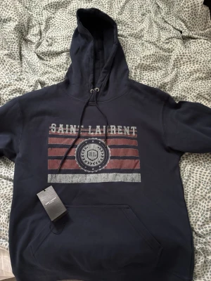 Mörkblå hoodie från Saint Laurent - Mörkblå hoodie från Saint Laurent med tryck framtill i vitt och rött, samt texten 'SAINT LAURENT' och ett emblem. Klassisk känguruficka och justerbar huva med snören. Perfekt för en avslappnad och trendig look.
