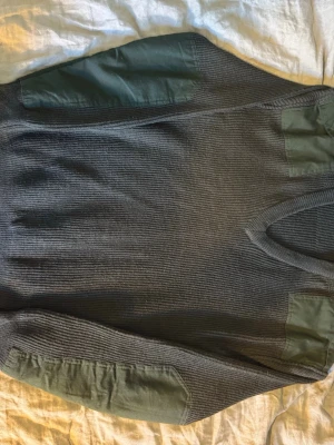 Militär mörkgrön stickad tröja med förstärkningar - Stickad tröja i mörkgrönt med förstärkta partier på axlar och armbågar i matchande tyg. Tröjan har rund halsringning och ribbstickade muddar. Perfekt för dig som gillar utility-stil och vill ha något slitstarkt och snyggt.