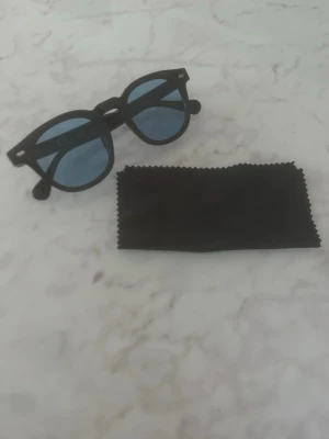 Svarta runda solglasögon - Snygga svarta solglasögon med rund form och blåtonade glas. Bågarna är i plast och glasögonen kommer med en svart putsduk. Perfekta för att höja din stil på soliga dagar. Helt nya alltså oanvända!