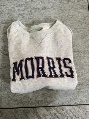 Grå sweatshirt från Morris - Snygg ljusgrå sweatshirt från Morris med stor mörkblå logga framtill. Tröjan har rund halsringning, ribbade muddar vid ärmslut och nederkant samt klassisk college-look. Perfekt för en avslappnad och trendig stil.