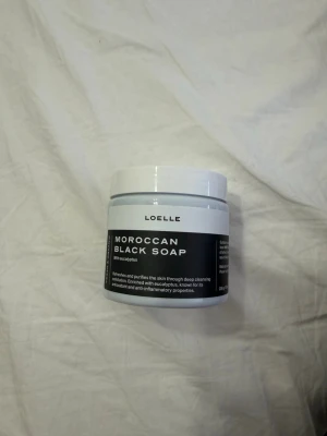 Loelle Moroccan Black Soap - Marockansk svart tvål från Loelle med eukalyptus i en rund vit plastburk. Tvålen rengör och återfuktar huden på djupet och är berikad med antioxidanter och antiinflammatoriska egenskaper. Perfekt för dig som vill ha en lyxig hudvårdsrutin.