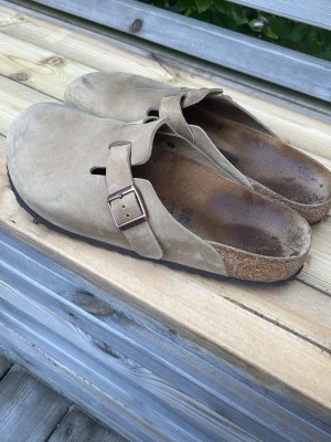 Beige Birkenstock tofflor  - Säljer ett par klassiska beige Birkenstock tofflor i mocka med justerbar rem och metallspänne. De har en ergonomisk fotbädd och en robust sula som ger bra komfort. Perfekta för dig som gillar stil och funktion.
