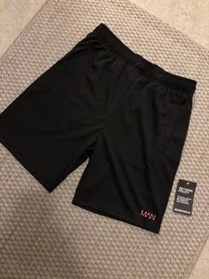 Svarta shorts från BoohooMAN -  Oanvänd helt ny. Svarta shorts från BoohooMAN med elastisk midja och diskret MAN-logga på ena benet. Klassisk rak modell i mjukt och lätt material, perfekta för en avslappnad stil. Passar till både chill och aktivitet.