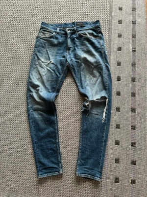 Blå slitna jeans med hål från Tiger of sweden - Säljer ett par blå jeans med slitningar och ett stort hål på höger knä. Jeansen har klassisk femficksdesign, normal midja och smal passform. Perfekta för en avslappnad och trendig streetstil. Materialet är denim i bomull.