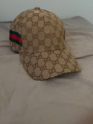 Beige keps - Stilren och exklusiv beige keps i det ikoniska GG-monogrammet. Kepsen har den klassiska beige/bruna färgkombinationen med gröna och röda webbränder på sidorna, vilket ger en tidlös och lyxig look. Tillverkad i Italien och utrustad med justerbart läderband baktill för optimal passform.
