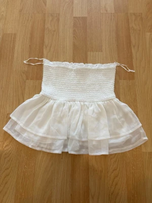 Vit bandeau tubtopp med volang Nelly - Vit bandeau tubtopp från Nelly med smockad överdel och dubbla lager volang i transparent tyg. Toppen är axelbandslös och har tunna knytband upptill. Perfekt för en luftig och trendig sommarstil. Har endast använt 1 gång🤍