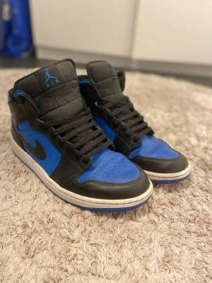 Air Jordan 1 High svart/blå sneakers - Säljer ett par Air Jordan 1 High sneakers i svart och blått läder med klassisk Nike swoosh på sidan och Jumpman-logga på tungan. Skorna har högt skaft, svarta skosnören och vit/blå sula. Perfekt för dig som gillar ikoniska och sportiga sneakers.