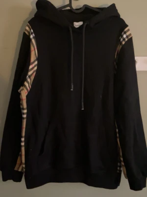 Burberry hoodie svart - Svart hoodie med klassiska beige, röda och svarta rutiga paneler längs ärmarna. Hoodien har huva med dragsko och en stor magficka framtill. Perfekt för dig som gillar streetwear och vill sticka ut med snygga detaljer.