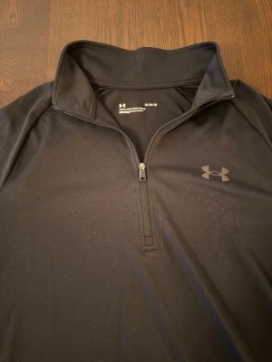 Under Armour träningströja  - Svart långärmad träningströja från Under Armour med halv dragkedja framtill och hög krage. Tillverkad i ett stretchigt funktionsmaterial som andas. Perfekt för löpning eller gym. Diskret logga på bröstet.