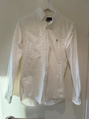 Vit skjorta från Ralph Lauren - Klassisk vit skjorta från Ralph Lauren med broderad logga på bröstet. Slim fit-modell med knappar framtill och krage. Tillverkad i mjuk bomull och har långa ärmar. Perfekt för en clean och stilren look.