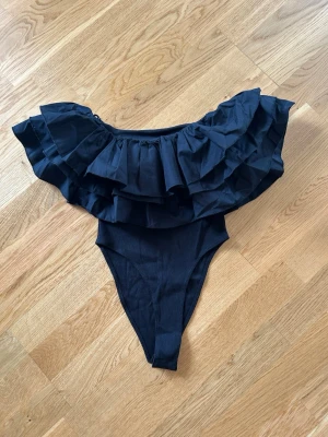 Ny trendig Svart body  med volanger från Zara  - Snygg svart body  med breda volanger över axlarna och off-shoulder design. Body  har en hög skärning och är tillverkad i ett stretchigt material som formar sig efter kroppen. Perfekt för dig som vill sticka ut 