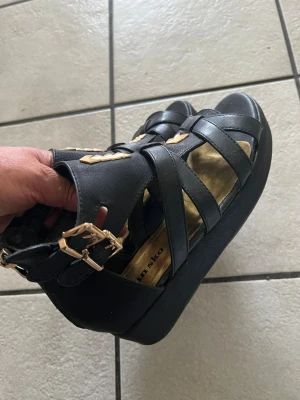 Svarta sandaler med nitar från Din Sko storlek 37 - Snygga svarta sandaler från Din Sko med breda remmar och guldfärgade nitar som dekoration. Sandalerna har en stadig platåsula och justerbart spänne vid ankeln. Tillverkade i konstläder med öppen tå och cool design.
