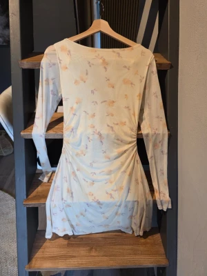 Ljusblommig gul  kort klänning med drapering i ryggen - Superfin kort klänning i ljus färg med subtilt blommigt mönster. Klänningen har lång ärm, draperad urringning och knyt i nacken. Tunt, lätt material som känns luftigt och perfekt för varmare dagar. Från Gina