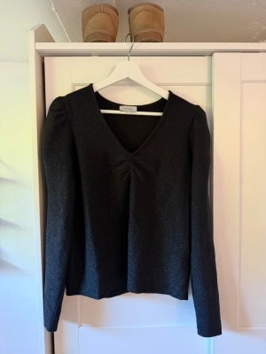 Svart glittrig topp - Snygg svart topp med glittrigt tyg, v-ringning och rynkdetalj framtill. Långärmad modell med lätt puff vid axlarna som ger en trendig siluett. Perfekt för dig som vill sticka ut lite extra.