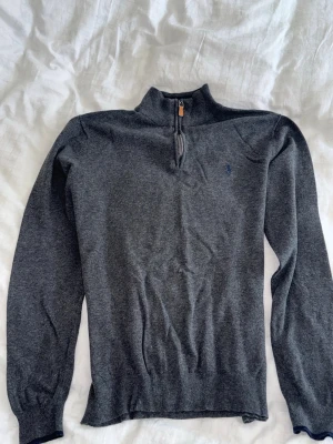 Grå halvzip från Polo Ralph Lauren - Mörkgrå tröja från Polo Ralph Lauren med halv dragkedja och liten broderad logga på bröstet. Tröjan har ribbade muddar vid ärmslut och nederkant samt hög krage. Perfekt för lager på lager och en clean look. XL på papper men L egentligen.