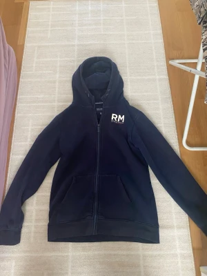 Mörkblå hoodie med dragkedja RM - Mörkblå hoodie med dragkedja framtill och vit RM-logga på bröstet. Klassisk huva och kängurufickor. Tillverkad i mjukt bomullsmaterial, perfekt för en avslappnad stil. Snygg och enkel design som passar till det mesta.