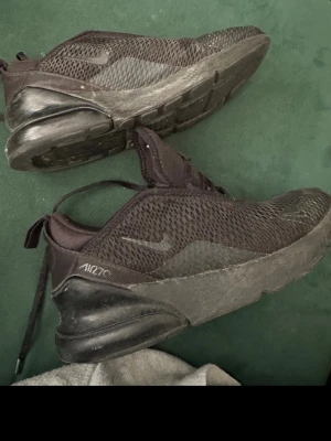 Nike Air Max 270 svarta sneakers - Säljer ett par helsvarta Nike Air Max 270 sneakers med mesh-ovandel och synlig Air-enhet i hälen. Skorna har en sportig look med diskret Nike-logga på sidan och snörning framtill. Perfekta för dig som gillar streetwear och bekväma skor.