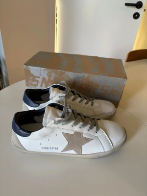Golden Goose vita sneakers med stjärna - Golden Goose sneakers i vitt läder med beige mockadetaljer och ikonisk stjärna på sidan. Svart hälparti och grå skosnören ger en cool kontrast. Snygg och tidlös design med lågt skaft och slitstark gummisula.
