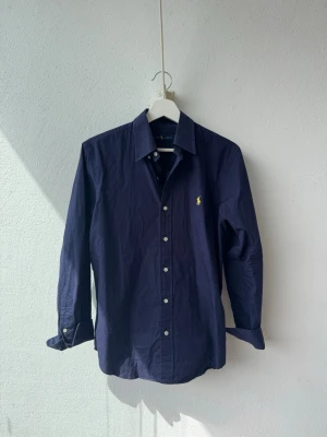 Mörkblå skjorta från Ralph Lauren - Klassisk mörkblå skjorta från Ralph Lauren med lång ärm och knappar framtill. Skjortan har en gul broderad logga på bröstet och är tillverkad i bomull. Perfekt för en stilren och tidlös look. Liten fläck vid ryggen men man märker den inte nästan alls och kanske far bort vid tvätt, vet inte