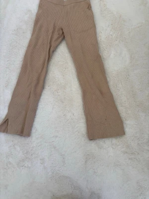 Byxor - Säljer ett par beige ribbade leggings med utsvängda ben och slitsar vid bensluten. De har hög midja och är gjorda i ett mjukt, stretchigt material som ger en skön passform. Perfekta för dig som gillar en trendig och avslappnad stil.
