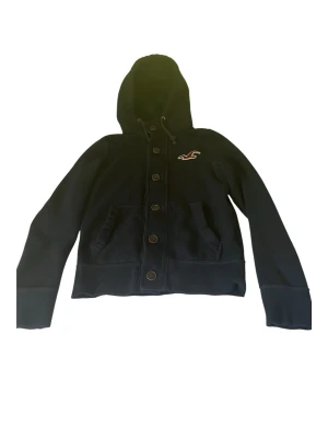 Svart hoodie från Hollister - Cool svart hoodie från Hollister med knappar framtill och två fickor. Den har en broderad logga på bröstet och en stor huva. Materialet är mjukt och bekvämt, perfekt för en avslappnad stil.