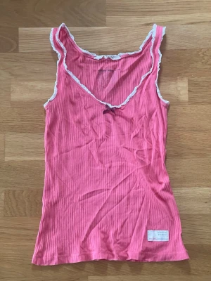 Rosa ribbad linnetopp med spetskant från Odd Molly i storlek S i fint skick - Söt rosa ribbad linnetopp med vit spetskant runt halsringningen och ärmhålen från Odd Molly. Storlek S. Liten rosett framtill och broderad detalj med texten 'RISE ABOVE' på ryggen. Perfekt för en avslappnad och trendig look.