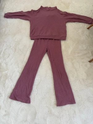 Rosa ribbat loungewear-set - Mysigt loungewear-set i ribbat material med tröja och byxor i en dämpad rosa nyans. Tröjan har hög hals och långa ärmar, byxorna har utsvängda ben. Perfekt för chill dagar hemma. Materialet känns mjukt och stretchigt.