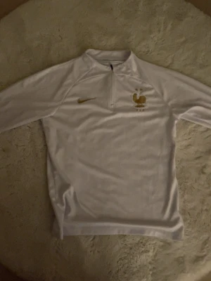 France football tracksuit - Snygg vit långärmad träningströja från Nike med Frankrikes landslagsemblem i guld på bröstet och guldfärgad Nike-logga. Tröjan har halv dragkedja framtill och är tillverkad i ett mjukt, sportigt material som passar perfekt till träning eller uppvärmning.