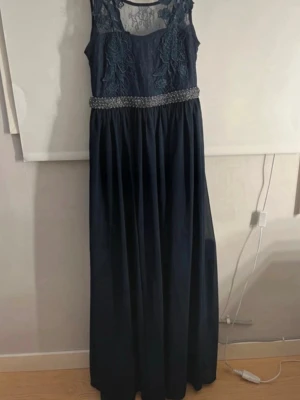 Marinblå långklänning med spets - Elegant marinblå långklänning med vackra spetsdetaljer och broderade blommor upptill. Klänningen har en dekorerad midja med pärlor och är ärmlös med transparent mesh upptill. Perfekt för bal eller festliga tillfällen.