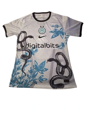 Inter Milan matchtröja Nike Thuram - Inter Milan matchtröja i vitt och ljusgrått med svarta detaljer, blå blommor och ormmotiv. Digitalbits-logga på bröstet, Nike-märke och Inter-emblem. Svart kant vid ärmar och hals. Nummer 9 och Thuram på ryggen. Perfekt för fotbollsfans!