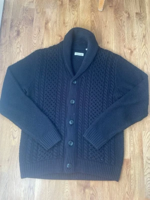 Svart stickad kofta Jack & Jones - Mysig svart stickad kofta från Jack & Jones med grovt kabelstickat mönster, sjalkrage och knäppning med fem knappar. Koftan har ribbade muddar vid ärmslut och nederkant. Perfekt för lager-på-lager och en stilren look.