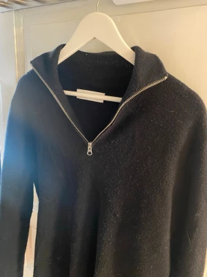 Svart halvzip-tröja från Samsøe Samsøe - Svart stickad tröja från Samsøe Samsøe med halvzip och hög krage. Tröjan har lång ärm och är tillverkad i ett mjukt, ulligt material som känns riktigt cozy. Perfekt för lager-på-lager och chill vibes.