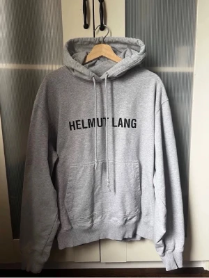 Grå Helmut Lang hoodie med tryck - Snygg grå hoodie från Helmut Lang med svart logotyptryck på bröstet. Klassisk modell med huva, dragsko och stor magficka. Tillverkad i mjukt bomullsmaterial och har ribbade muddar vid ärmslut och midja. Perfekt för en avslappnad streetstyle-look.