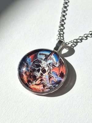 Halsband med dödskalle och svärd - halsband med metallkedja och hänge med motiv av en dödskalle, svärd och drake i 2D, färgerna svart, blått, rött och vitt. Hänget har en glansig yta och en cool fantasy-inspirerad design. Perfekt för dig som gillar unika och tuffa accessoarer.
